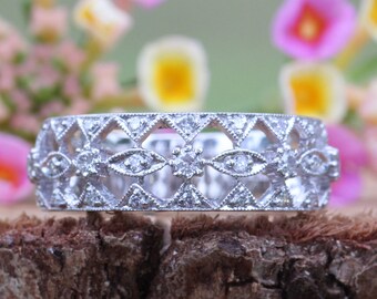 May include: A delicate silver ring with a filigree design and small diamonds set in the band. The ring is resting on a piece of wood with pink flowers in the background.