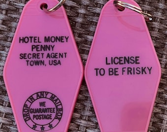 Può includere: Due portachiavi di motel rosa con testo nero. Un portachiavi dice "Hotel Money Penny Secret Agent Town, USA" e ha un cerchio nero con il testo "Drop In Any Mailbox We Guarantee Postage". L'altro portachiavi dice "License To Be Frisky".