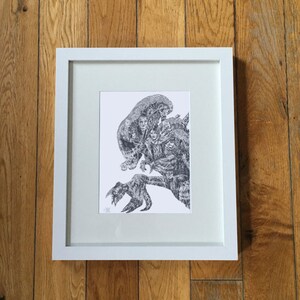 May include: A framed black and white print showing a detailed illustration of a creature with multiple faces and a clawed hand. The artwork is set against a white background within a white frame, displayed on a wooden surface.