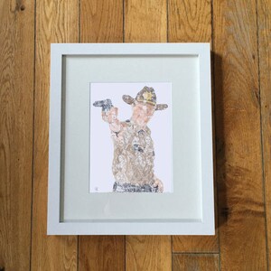 May include: Framed art print featuring a figure in a sheriff's hat holding a gun. The figure is composed of a collage of faces in shades of brown and tan. The white framed print is displayed on a wooden surface.