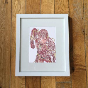 May include: A framed print featuring a red and gold illustration of a figure composed of smaller figures. The artwork is set against a white background within a white frame, displayed on a wooden surface.
