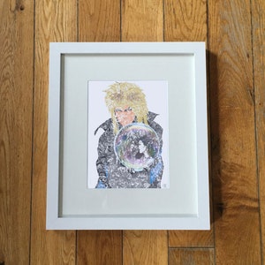 May include: A framed art print featuring a portrait of a person with blonde hair and a patterned jacket, holding a crystal ball. The crystal ball contains an image of two people. The frame is white, and the artwork is displayed on a wooden surface.