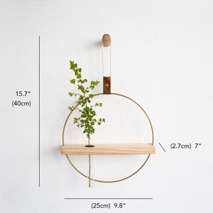 May include: A gold metal ring with a wooden shelf attached. The ring is hanging from a brown leather strap and a wooden hook. A small plant is in a glass tube on the shelf. The ring is 9.8 inches in diameter and the shelf is 7 inches long.