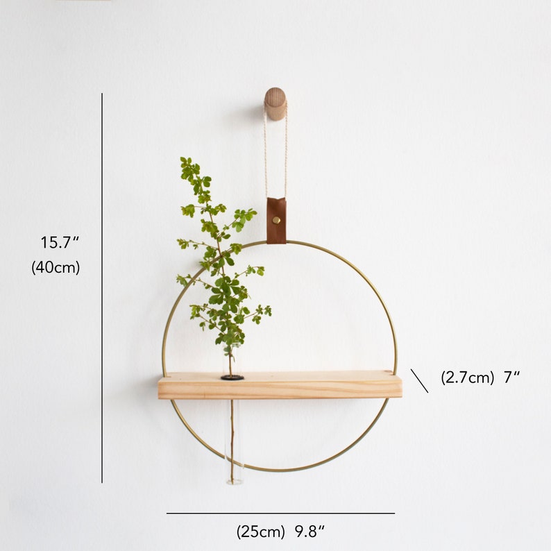 May include: A gold metal ring with a wooden shelf attached. The ring is hanging from a brown leather strap and a wooden hook. A small plant is in a glass tube on the shelf. The ring is 9.8 inches in diameter and the shelf is 7 inches long.