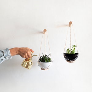May include: Two hanging planters with plants, one white and one black, with a gold watering can in the foreground.