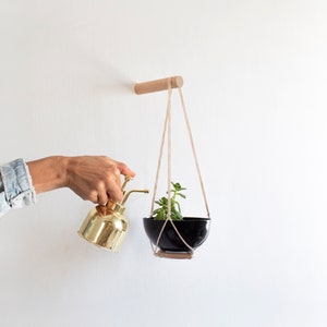 Pode incluir: Uma pessoa está regando uma planta suspensa com um regador de latão. A planta está em um vaso preto e está pendurada em uma cavilha de madeira com cordão de macramé.
