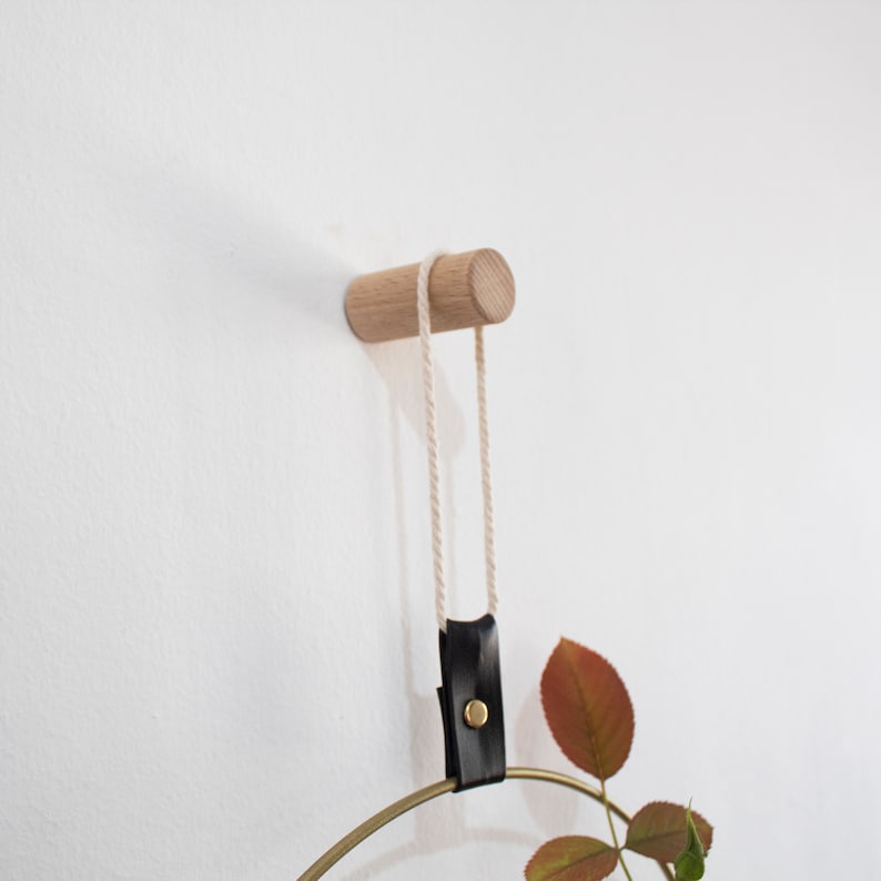 May include: A wooden wall hook with a natural finish and a leather strap hanging from it. The strap is attached to a gold metal hoop with a plant hanging from it.