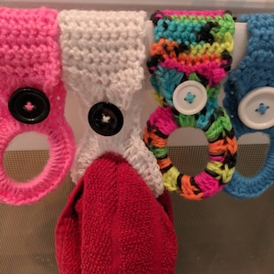 Peut inclure: Quatre porte-serviettes en crochet avec boutons. Les porte-serviettes sont en rose, blanc, arc-en-ciel et bleu. Une serviette rouge est accrochée au porte-serviettes blanc.