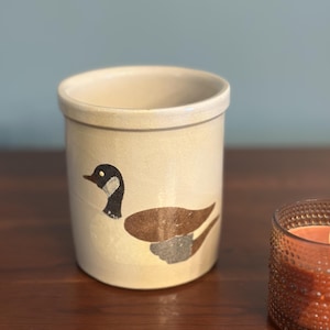May include: A cylindrical ceramic crock with a cream-colored glaze featuring a painted graphic of a Canada goose. The goose has a black head and neck with a white cheek patch and brown wings. The vessel has a thick rim and a flat base.