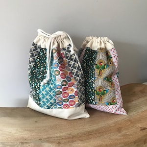Pode incluir: Duas sacolas com cordão com designs de patchwork coloridos. Uma sacola apresenta um padrão turquesa, verde e vermelho, enquanto a outra tem um design rosa, verde e amarelo. Ambas as sacolas têm uma parte superior e inferior creme.