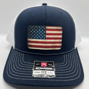 May include: A navy blue and white trucker hat with a distressed American flag patch. The hat features a curved brim, white mesh back, and the text "RICHARDSON ORIGINAL 112".
