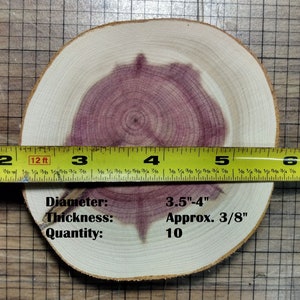 May include: A close-up of a round wooden slice with a purple heartwood pattern. The slice is approximately 3.5 to 4 inches in diameter and 3/8 inch thick. A measuring tape is placed across the slice, showing the diameter. The text "Diameter: 3.5"-4"" and "Thickness: Approx. 3/8"" is written below the slice. The text "Quantity: 10" is written below the thickness.
