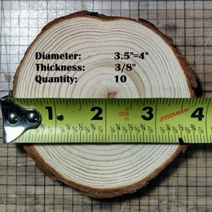 May include: A close-up of a wood slice with a diameter of 3.5 inches or 4 inches and a thickness of 3/8 inch. The wood slice is light brown in color and has a natural wood grain pattern. A measuring tape is placed on the wood slice, showing the measurement of 1 inch to 4 inches. The text "Diameter: 3.5"=4"", "Thickness: 3/8"", and "Quantity: 10" is written on the wood slice.