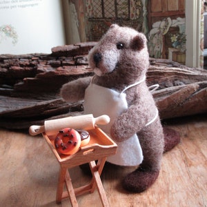 May include: A brown felt marmot wearing a white apron stands next to a small wooden table. The table has a rolling pin and two doughnuts on it. One doughnut is chocolate with sprinkles and the other is a plain glazed doughnut.