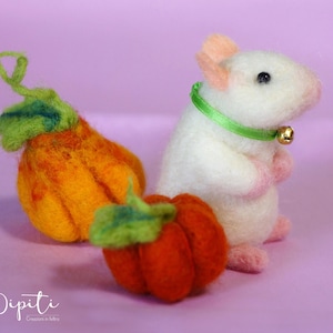 May include: A white felt mouse wearing a green ribbon and a gold bell, sitting next to two orange felt pumpkins with green stems.