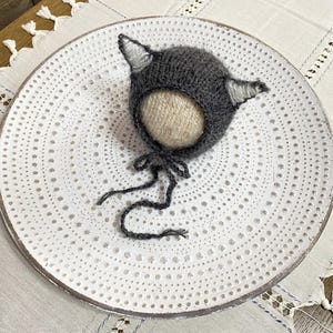 Può includere: Cappellino per neonato di lana grigia con orecchie a punta. Il cappello è su un piatto bianco con un motivo a pois.