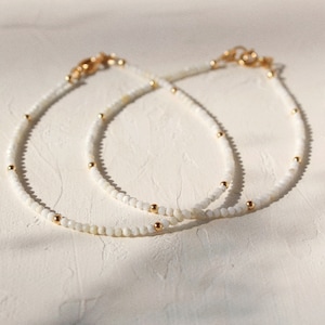 Peut inclure: Deux colliers délicats avec des perles blanches et des accents dorés. Les colliers sont superposés sur une surface blanche.