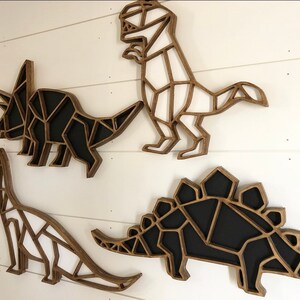 May include: Four wooden dinosaur wall decorations in a geometric style. The dinosaurs are a triceratops, a stegosaurus, a brachiosaurus, and a tyrannosaurus rex. The dinosaurs are painted in black and white.