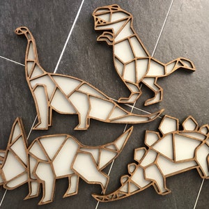 May include: Four wooden dinosaur wall decorations in a geometric style. The dinosaurs are a stegosaurus, a triceratops, a brachiosaurus, and a tyrannosaurus rex. The dinosaurs are all white with brown outlines.