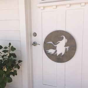 May include: A white front door with a wooden circle cutout of a witch flying on a broomstick. The door has a silver door handle and a silver door knob. A green plant with yellow flowers is to the left of the door.