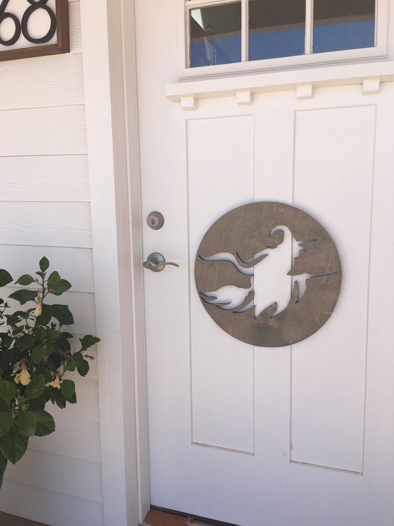 May include: A white front door with a wooden circle cutout of a witch flying on a broomstick. The door has a silver door handle and a silver door knob. A green plant with yellow flowers is to the left of the door.