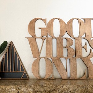 May include: Wooden sign with the words "Good Vibes Only" in a modern font. The sign is a light brown color and has a rustic look. It is sitting on a shelf with a small cactus plant and a black triangle-shaped object in the background.