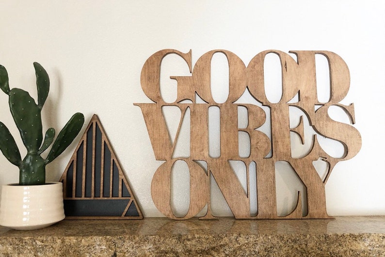 May include: Wooden sign with the words "Good Vibes Only" in a modern font. The sign is a light brown color and has a rustic look. It is sitting on a shelf with a small cactus plant and a black triangle-shaped object in the background.