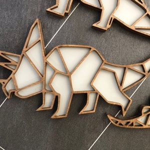 May include: A wooden cutout of a triceratops dinosaur, painted white, with a geometric design. The dinosaur is facing left and has a long horn on its nose and two shorter horns above its eyes.