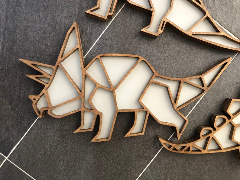 May include: A wooden cutout of a triceratops dinosaur, painted white, with a geometric design. The dinosaur is facing left and has a long horn on its nose and two shorter horns above its eyes.