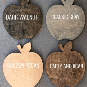 May include: Four wooden apple-shaped signs with different wood stains. The signs are labeled "Dark Walnut", "Classic Gray", "Golden Pecan", and "Early American".