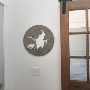May include: A wooden wall decor featuring a silhouette of a witch flying on a broomstick. The silhouette is cut out of the wood, creating a negative space effect. The wood is a light brown color.