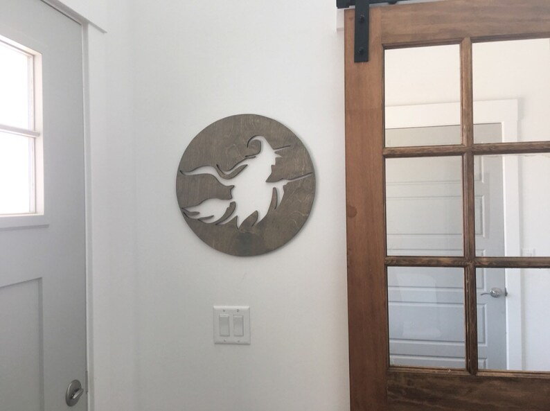 May include: A wooden wall decor featuring a silhouette of a witch flying on a broomstick. The silhouette is cut out of the wood, creating a negative space effect. The wood is a light brown color.