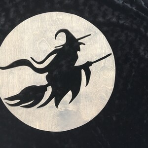 May include: A black silhouette of a witch flying on a broomstick against a white moon background. The witch is wearing a pointed hat and has long flowing hair. The moon is a circle with a rough wood grain texture.