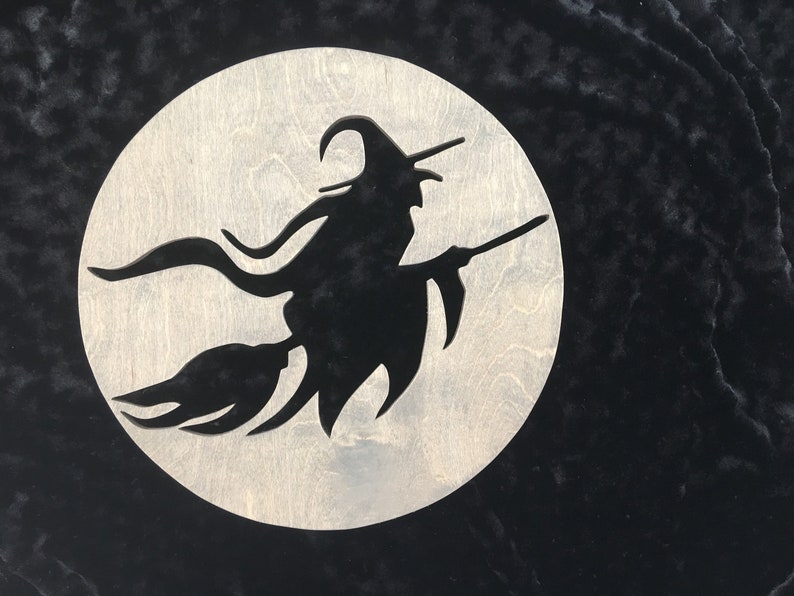 May include: A black silhouette of a witch flying on a broomstick against a white moon background. The witch is wearing a pointed hat and has long flowing hair. The moon is a circle with a rough wood grain texture.