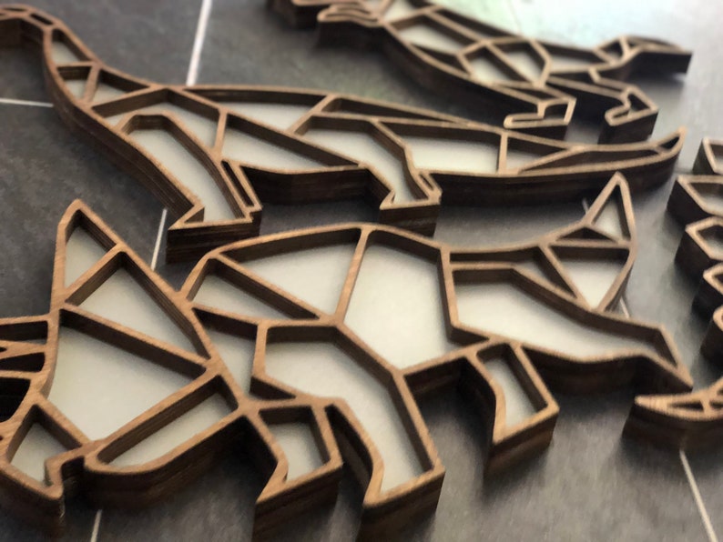 May include: A close-up of a set of wooden dinosaur ornaments. The dinosaurs are made of a light brown wood and have a geometric design. The ornaments are stacked on top of each other.