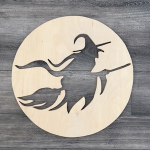 May include: A wooden cutout of a witch flying on a broomstick, set against a plain wooden background. The witch is silhouetted against a circular piece of wood.