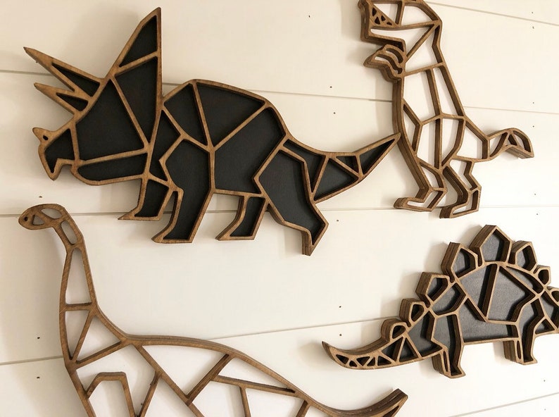 May include: Four wooden dinosaur wall decorations in a geometric style. The dinosaurs are a triceratops, a stegosaurus, a brachiosaurus, and a tyrannosaurus rex. The dinosaurs are all black and white.