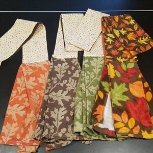 May include: A collection of decorative kitchen towels with various fall-themed patterns. The towels feature designs of autumn leaves and oak leaves in shades of orange, brown, and green. The tops of the towels have a dotted pattern.