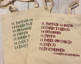 May include: Two cream-colored tote bags with text in green and burgundy. The text on the bags includes phrases like "El Justo de la Noche" and "Leyendas Salvadoreñas." The bags have long handles and are made of a light-colored fabric.