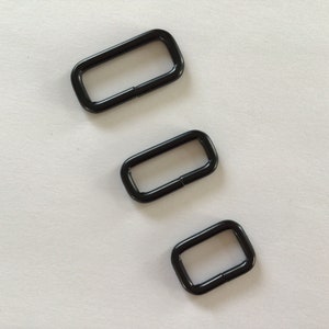 Peut inclure: Trois anneaux en D en métal noir de différentes tailles. Les anneaux sont sur un fond blanc.