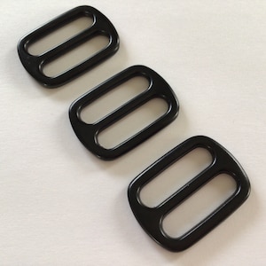 May include: Three black plastic slider buckles. The buckles are rectangular with two oval-shaped holes in the centre.