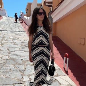 Puede incluir: Un conjunto de dos piezas de crochet en blanco y negro, que consta de un top sin mangas y una falda larga con dobladillo con flecos. El conjunto tiene un patr&oacute;n de chevr&oacute;n. La persona lleva sandalias negras y un bolso negro.