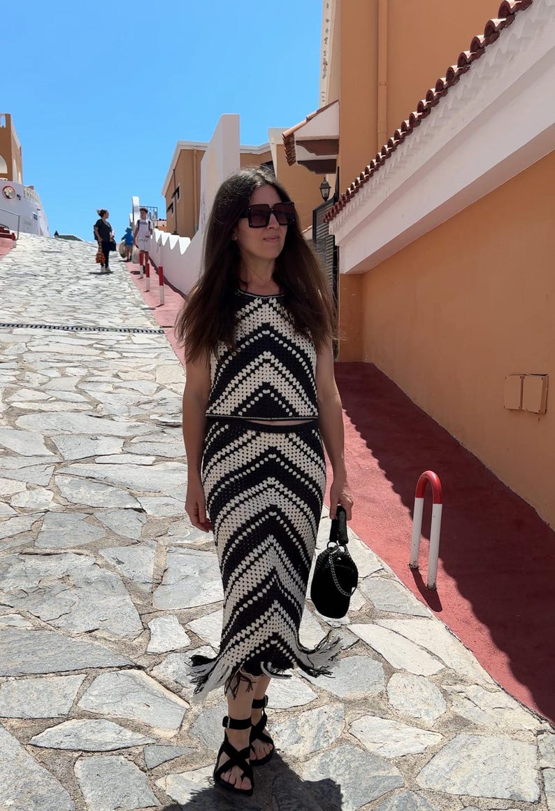 Puede incluir: Un conjunto de dos piezas de crochet en blanco y negro, que consta de un top sin mangas y una falda larga con dobladillo con flecos. El conjunto tiene un patr&oacute;n de chevr&oacute;n. La persona lleva sandalias negras y un bolso negro.