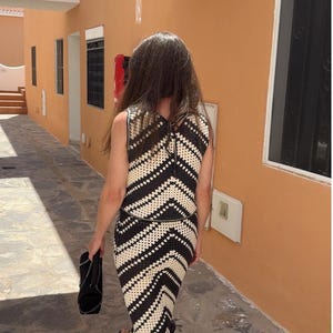 Puede incluir: Conjunto de dos piezas de ganchillo en blanco y negro, con un top sin mangas y una falda con flecos. El conjunto se lleva con sandalias negras y un bolso de mano negro. El fondo muestra un edificio naranja.