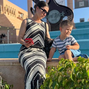 Puede incluir: Una mujer con un vestido de ganchillo blanco y negro con flecos, sandalias negras y gafas de sol. Un ni&ntilde;o peque&ntilde;o con una camisa a rayas y pantalones cortos azules est&aacute; cerca. El fondo muestra un letrero de Hard Rock Cafe y una guitarra grande.