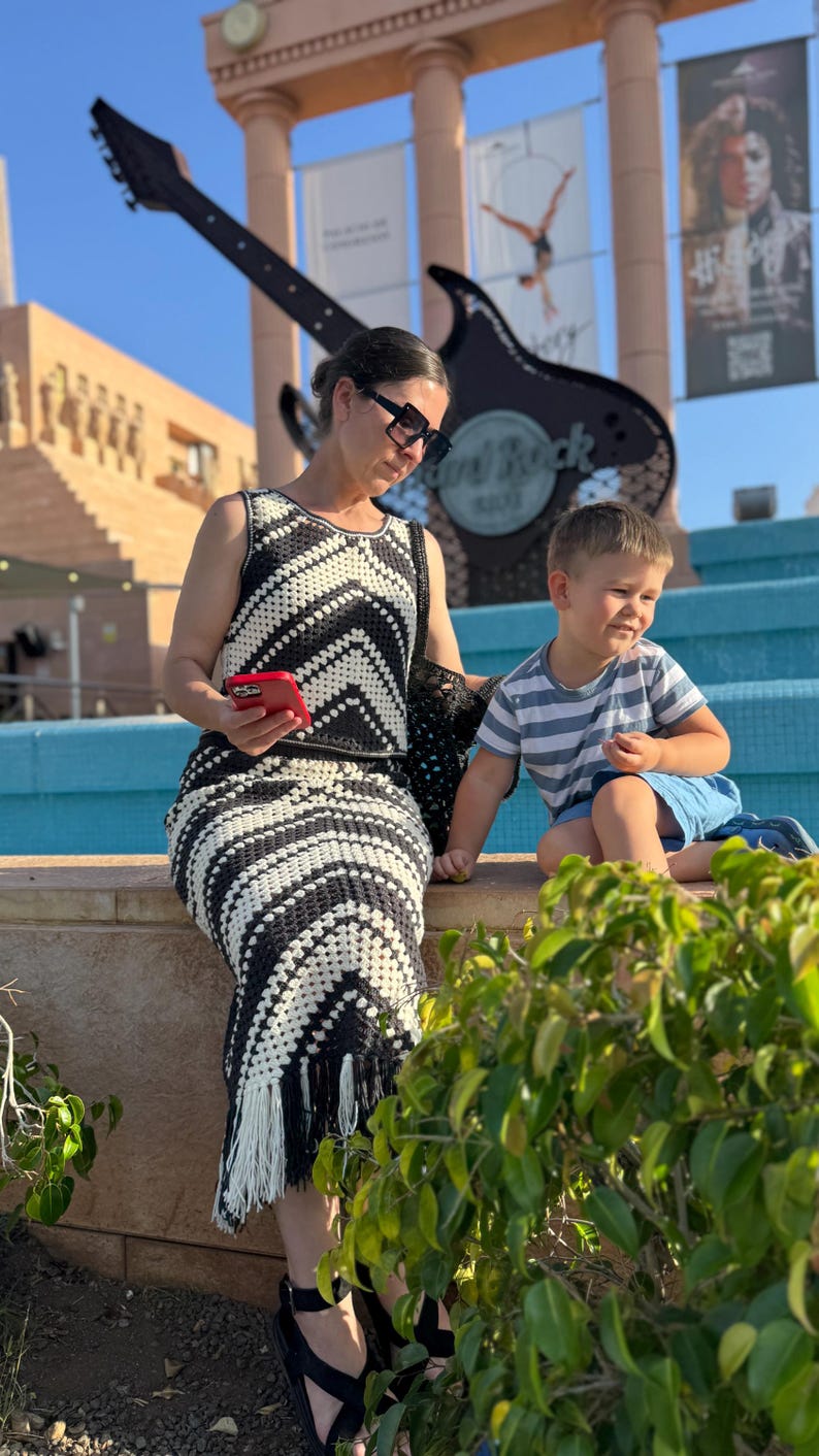 Puede incluir: Una mujer con un vestido de ganchillo blanco y negro con flecos, sandalias negras y gafas de sol. Un ni&ntilde;o peque&ntilde;o con una camisa a rayas y pantalones cortos azules est&aacute; cerca. El fondo muestra un letrero de Hard Rock Cafe y una guitarra grande.