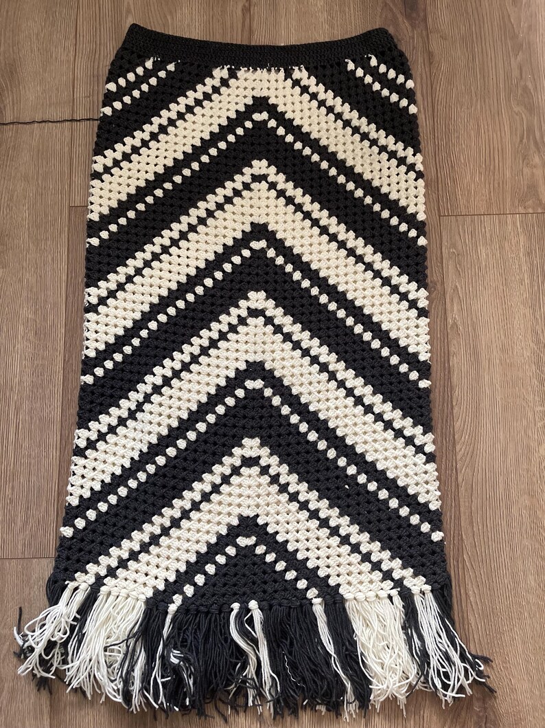 Puede incluir: Falda de crochet negra y crema con un patr&oacute;n de chevr&oacute;n. La falda tiene una cintura alta y una silueta larga y recta. El borde inferior est&aacute; rematado con una franja de hilo negro y crema.