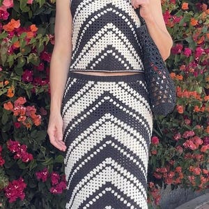 Puede incluir: Conjunto de dos piezas de crochet en blanco y negro, con un top sin mangas y una falda larga con flecos. El patr&oacute;n geom&eacute;trico es un dise&ntilde;o de chevr&oacute;n. Se lleva un bolso de malla negro.