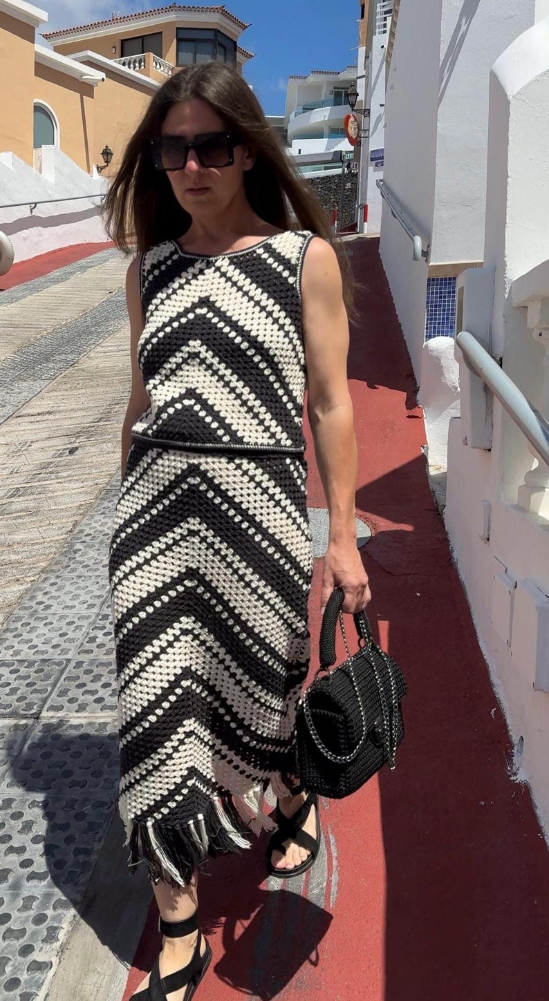 Puede incluir: Vestido de crochet en blanco y negro con un patr&oacute;n de chevron. El vestido sin mangas tiene un cintur&oacute;n a juego y flecos en el dobladillo. Un bolso negro con una correa de cadena y sandalias negras completan el conjunto.