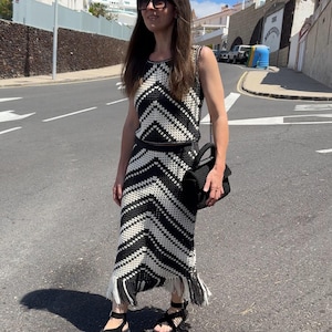 Puede incluir: Conjunto de dos piezas de crochet en blanco y negro, con un top sin mangas y una falda larga con flecos. El conjunto se combina con sandalias negras y un bolso de mano negro. El conjunto tiene un patr&oacute;n de chevr&oacute;n.